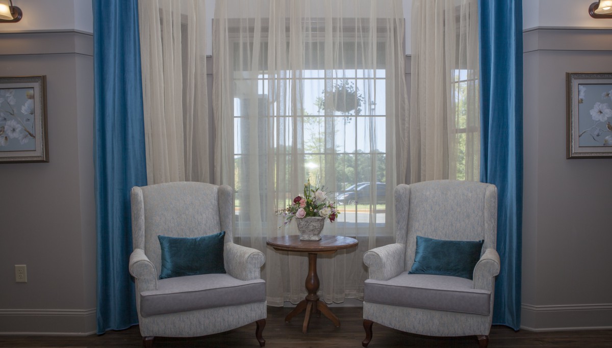 A cozy living room setting with two armchairs and a small table adorned with flowers, complemented by sheer curtains.