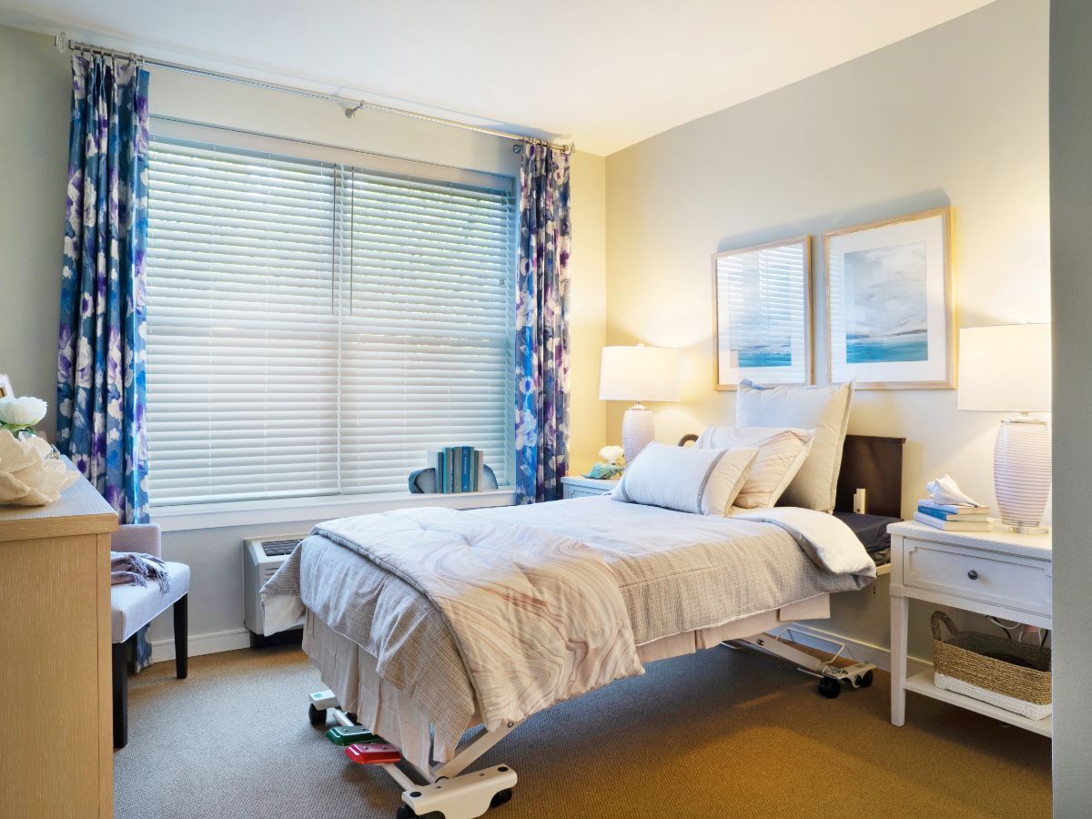 A cozy hospital room with a neatly made bed, window with floral curtains, and framed artwork on the wall.