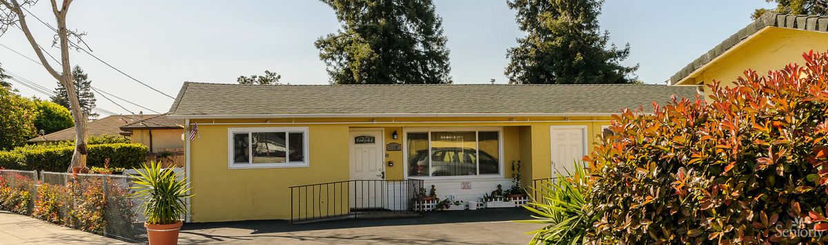 A yellow single-story house with a front porch and driveway, set against a backdrop of trees.