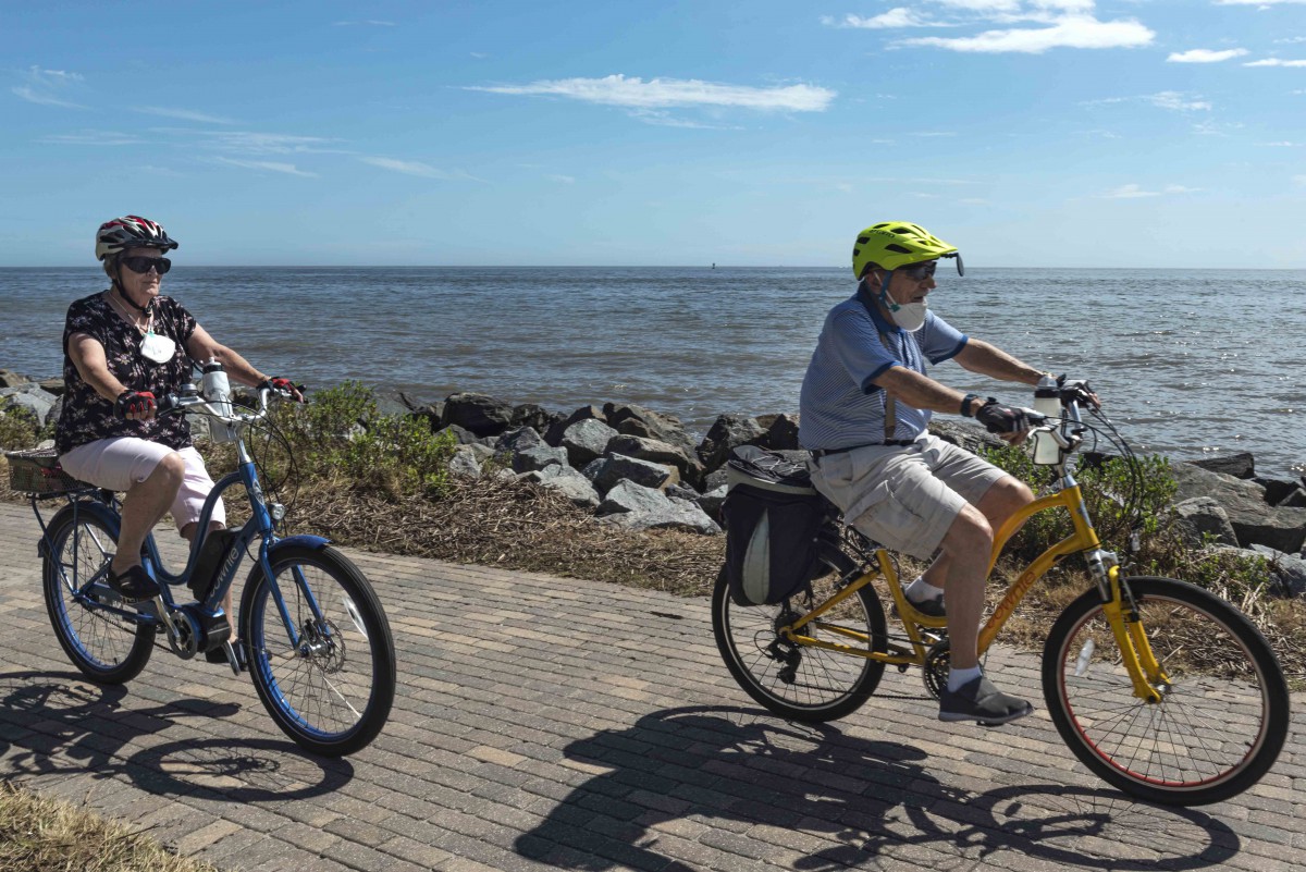Two people biking along a scenic boardwalk by the ocean on a sunny day.