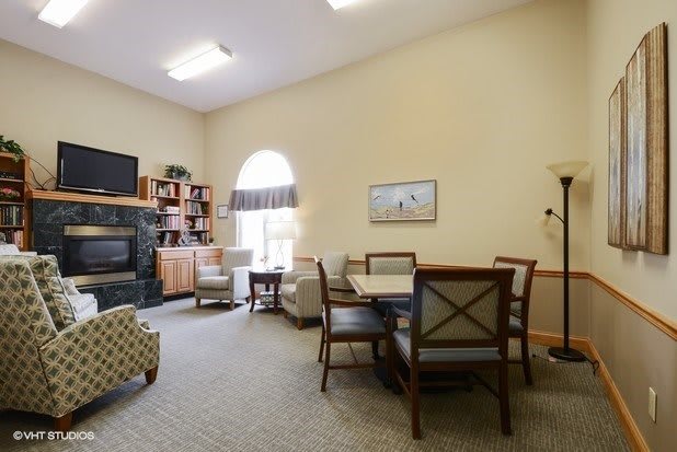 A cozy living room with comfortable chairs, a small table, bookshelves, and a television, illuminated by natural light from an arched window.