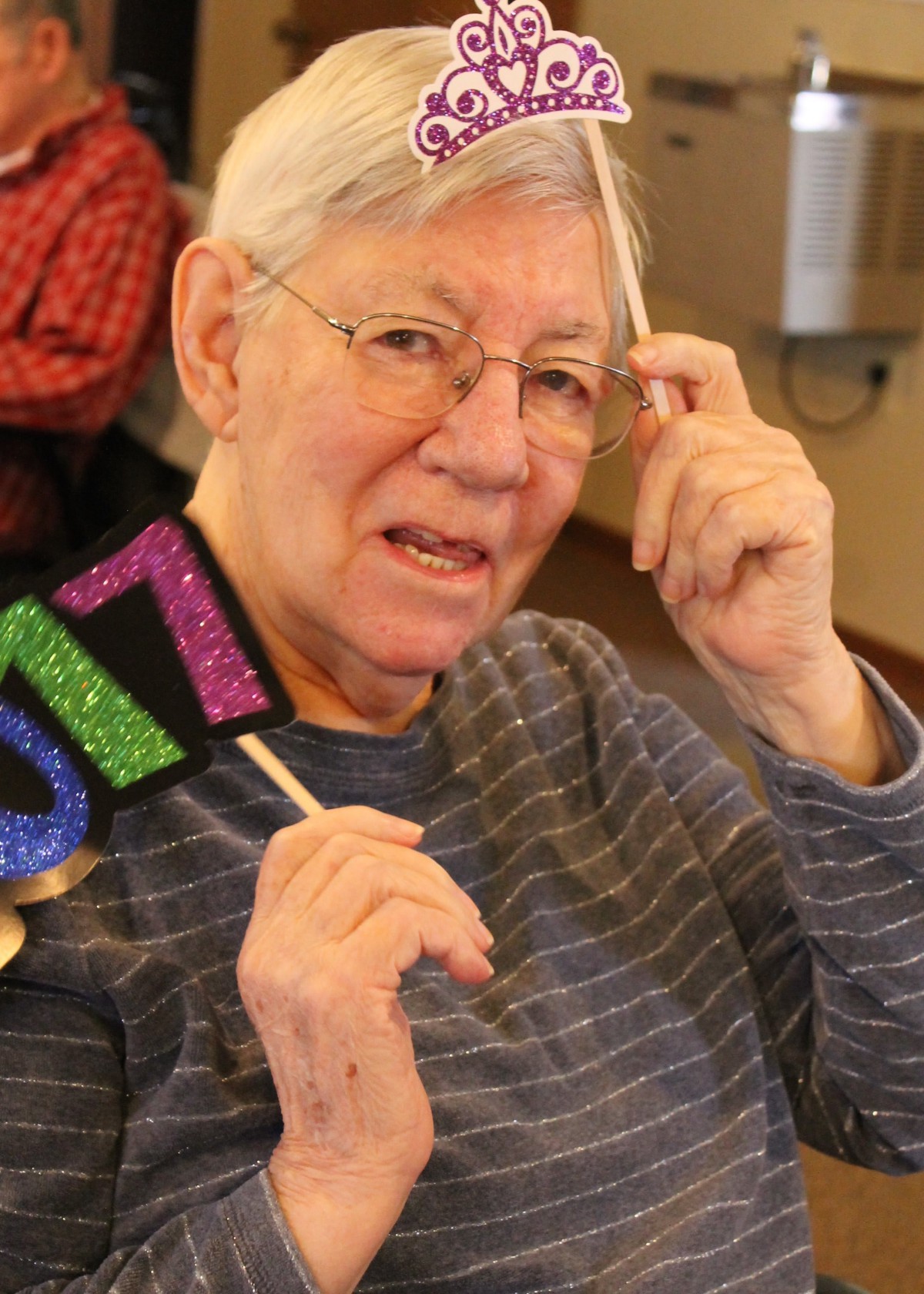 An elderly person wearing glasses holds a colorful '2017' sign while sitting indoors, possibly at a celebration.