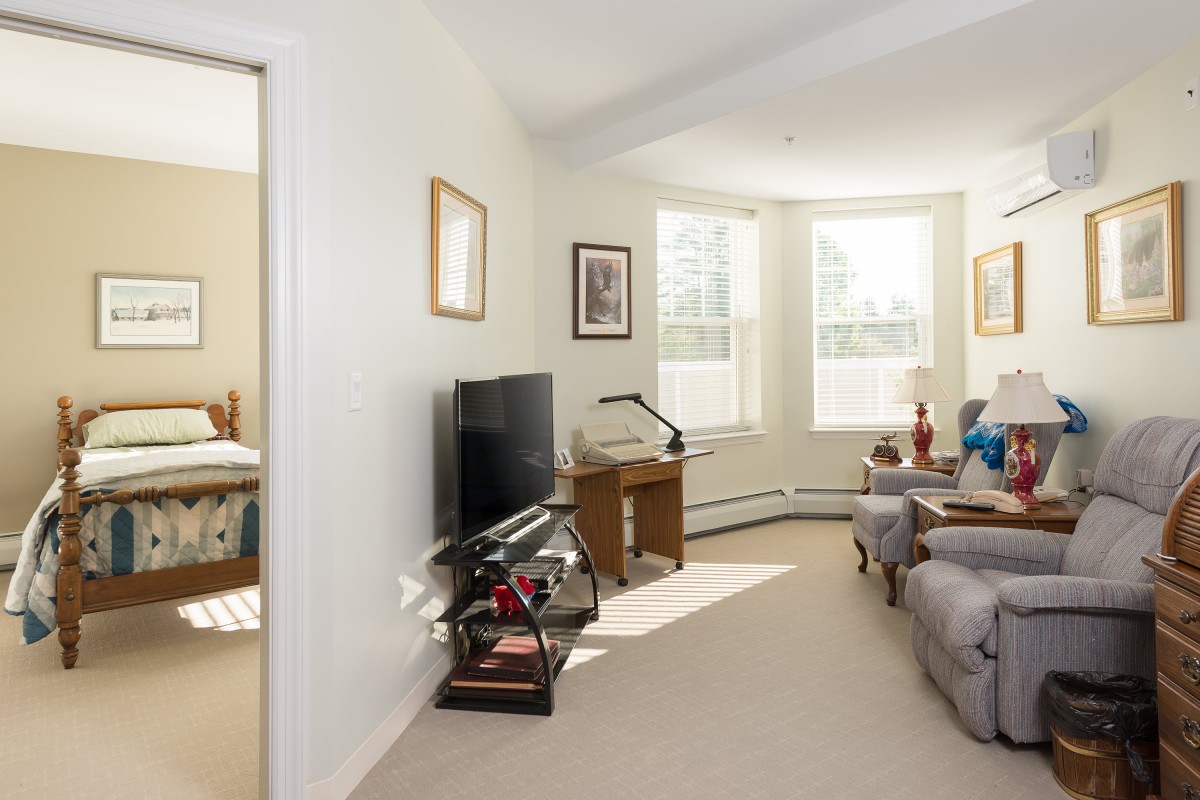 A cozy living room with natural light, featuring comfortable seating, a television, and a small desk.