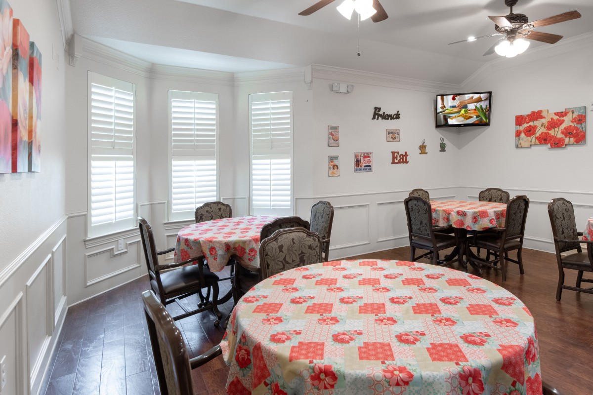 A cozy dining area with round tables covered in patterned tablecloths, surrounded by chairs and adorned with wall decorations. A television is mounted in the corner.