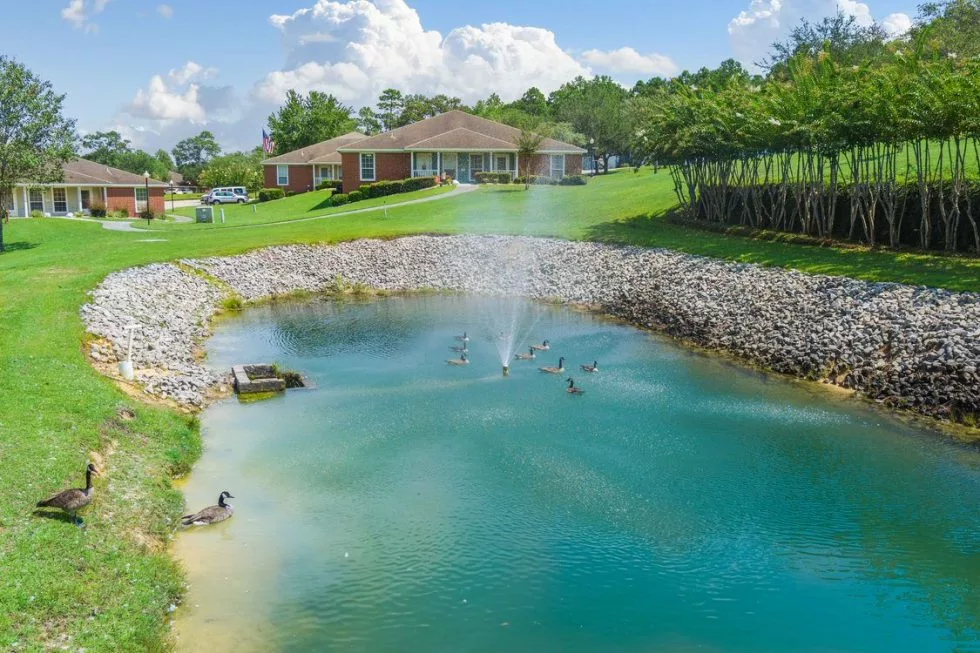 A serene landscape featuring a pond with a fountain in front of a house surrounded by grass and trees.
