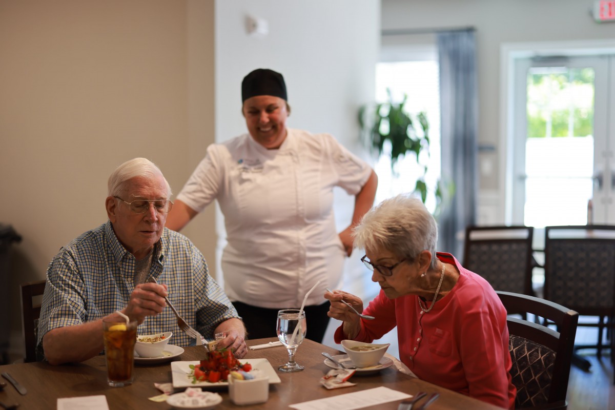 A chef engaging with two seniors while they enjoy a meal at a dining table.
