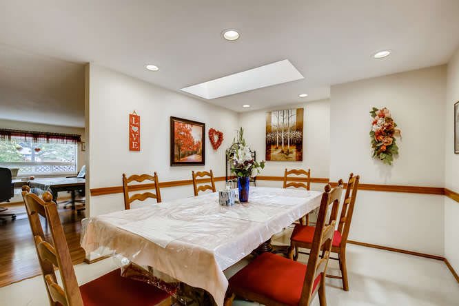 A well-lit dining room featuring a table with a white tablecloth, surrounded by wooden chairs with red cushions. Walls are adorned with artwork and decorations.