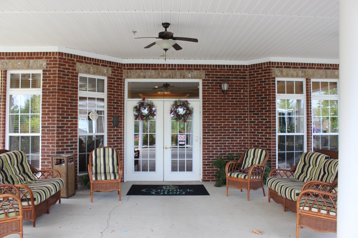A cozy porch with brick walls and comfortable armchairs, featuring French doors adorned with wreaths and a ceiling fan overhead.