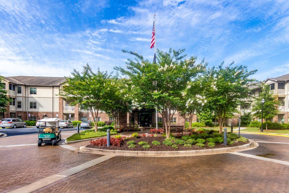 A well-landscaped residential entrance with trees and paving, featuring an American flag flying above.