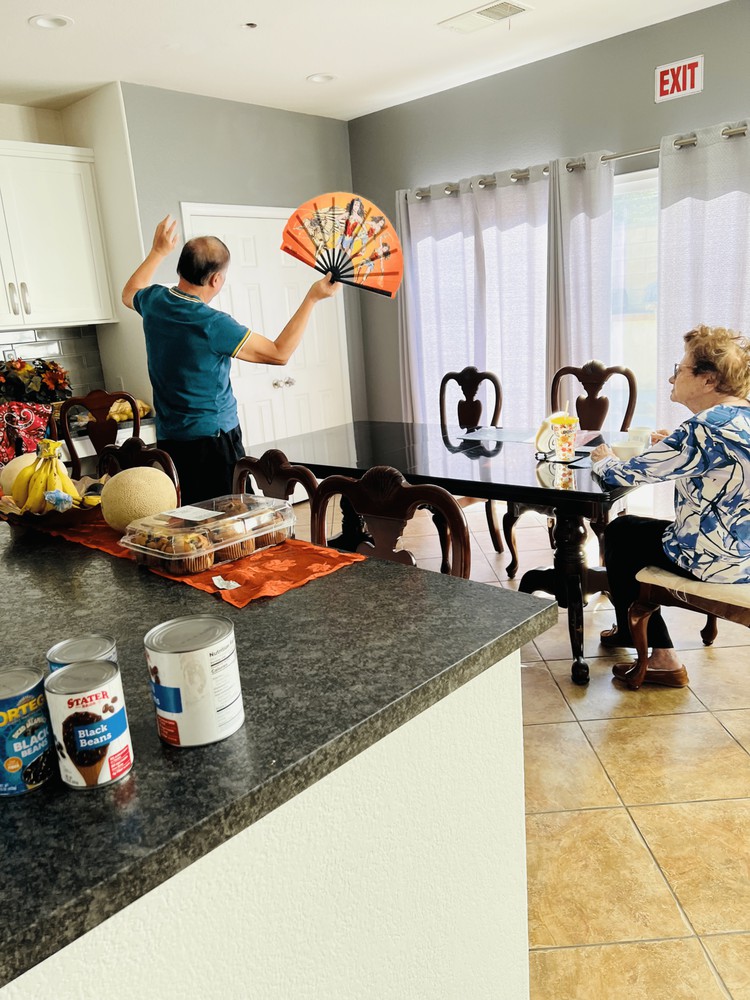 Two people in a kitchen, one holding a decorative fan while another sits at the dining table.
