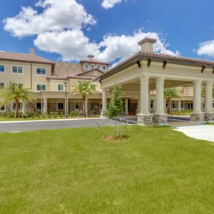 A large residential building with a covered entrance and manicured lawn under a bright blue sky with clouds.