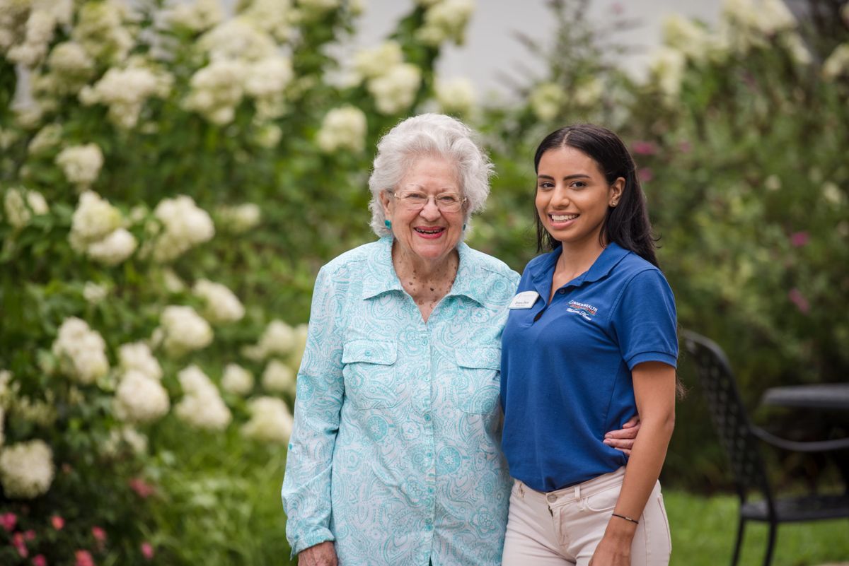 An elderly woman and a young woman smiling in a garden full of flowers.