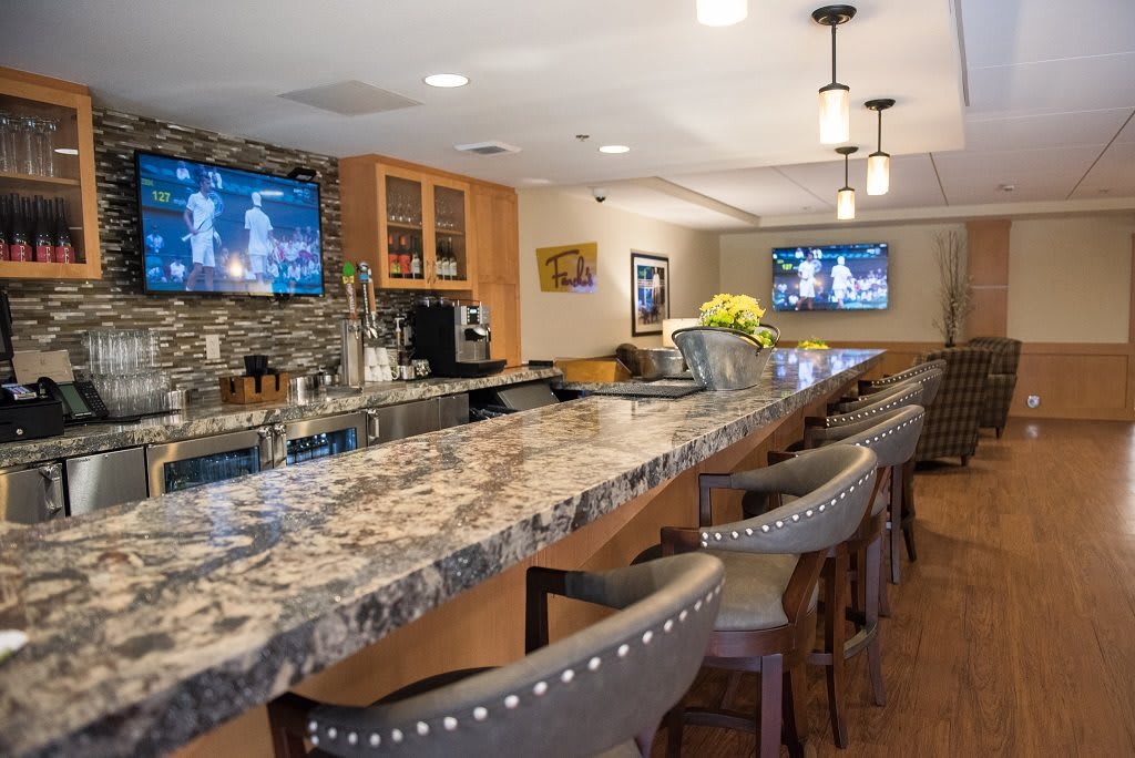 A modern indoor bar with a granite countertop, stylish chairs, and televisions.