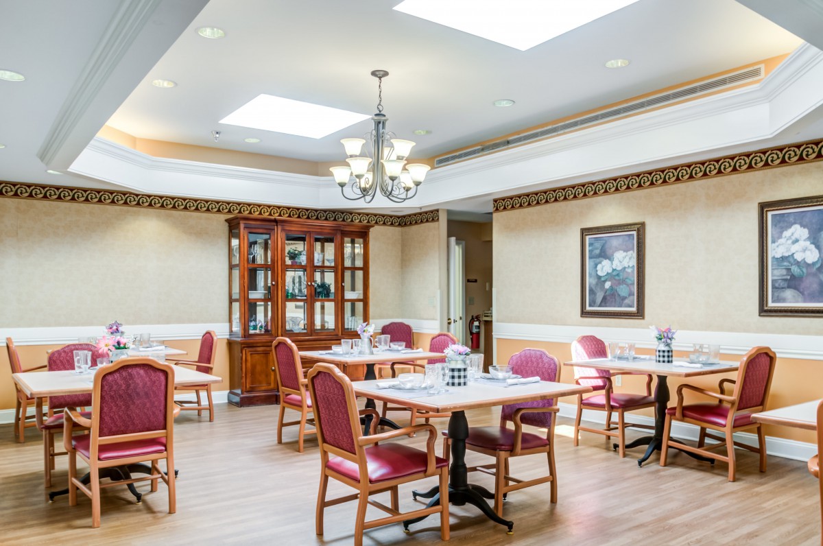 A well-lit dining room with elegant tables and chairs, featuring a chandelier and a wooden cabinet.