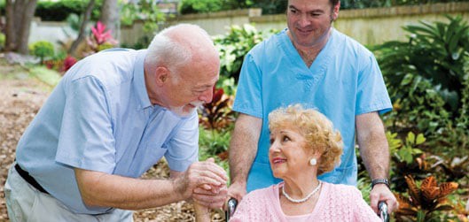 A caregiver assisting an elderly woman in a wheelchair, with another person engaging with them outdoors.