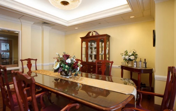A formal dining room with a polished wooden table, surrounded by chairs, decorated with floral arrangements and a china cabinet in the background.