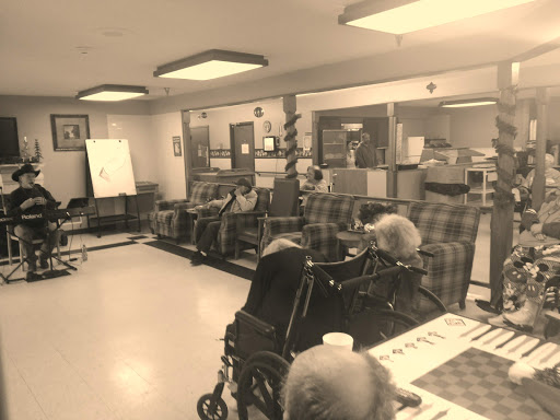A community room in a healthcare facility with elderly individuals seated and engaged in conversation.