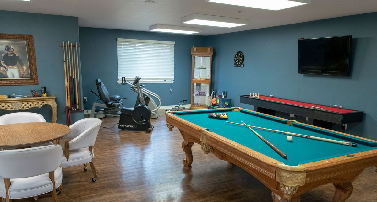 A spacious game room featuring a pool table, exercise bike, and shuffleboard on wood flooring against blue walls.