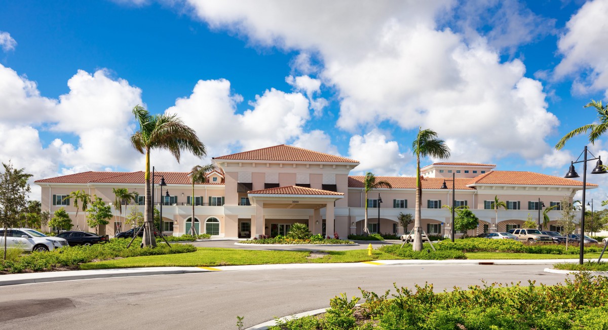 A large building with a red-tiled roof surrounded by palm trees and set against a backdrop of blue sky and clouds.