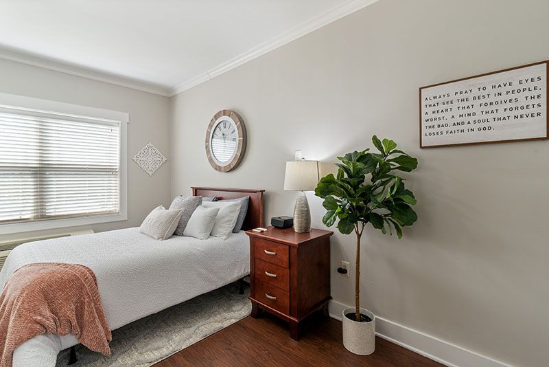 A cozy bedroom featuring a bed with white linens, a wooden nightstand with a lamp, and a decorative plant next to the wall clock.