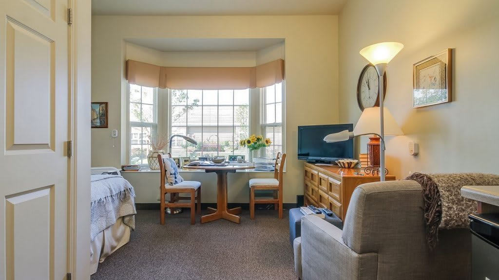 A cozy room with a large window, featuring a desk, chairs, bed, and television.