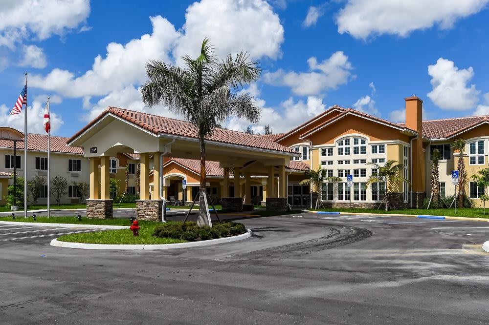A modern residential building with palm trees and a landscaped driveway under a partly cloudy sky.