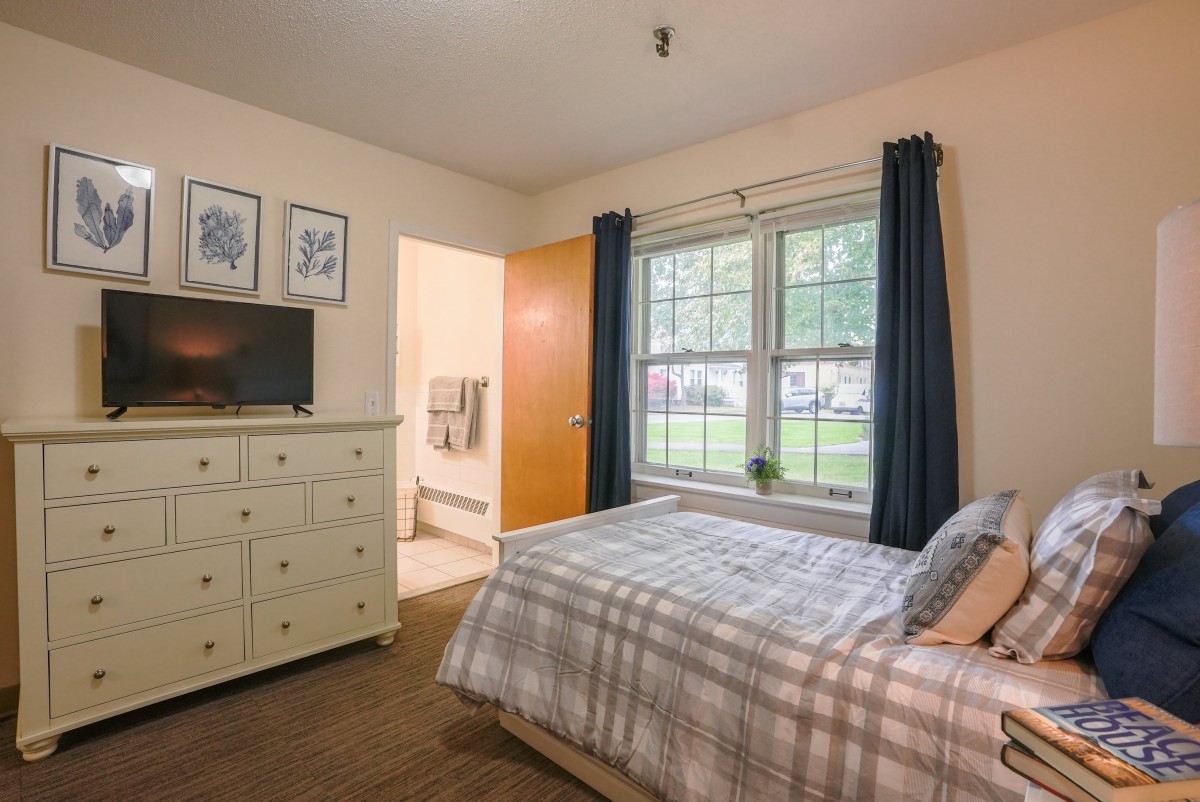 A cozy bedroom with a bed, dresser, and window, featuring soft lighting and neutral decor.