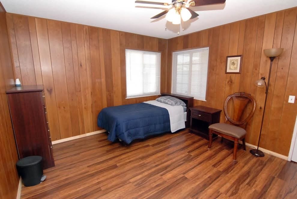 A cozy bedroom with wood paneling, featuring a single bed with blue bedding, a hardwood floor, and a ceiling fan.