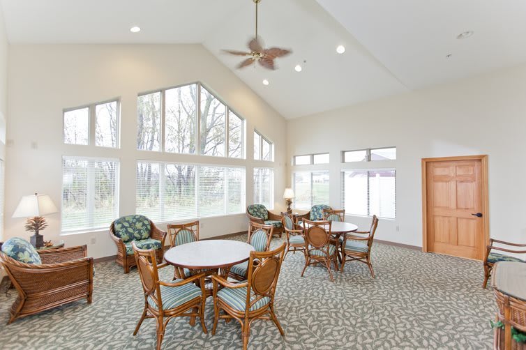 A spacious room with large windows, tables, and chairs, illuminated by natural light.