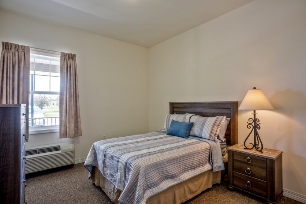A cozy bedroom with a neatly made bed, nightstand, and a lamp by the window.
