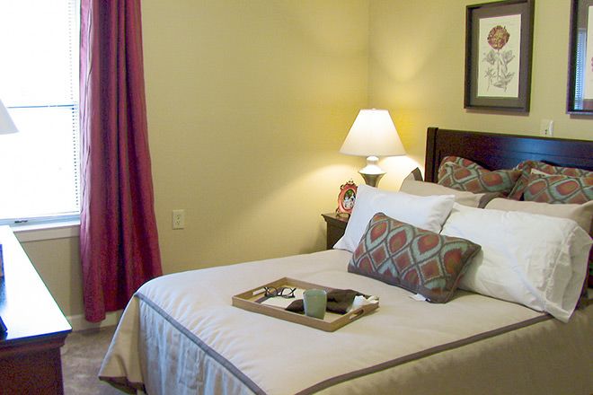 A cozy bedroom with a neatly made bed, adorned with pillows and a decorative tray, lit by a warm lamp.