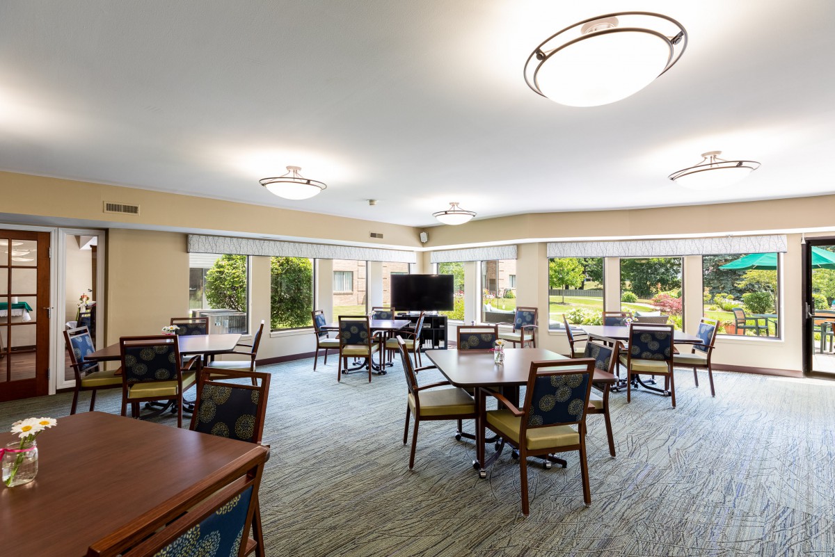 A spacious and well-lit room with multiple tables and chairs arranged for seating, featuring large windows and a television.