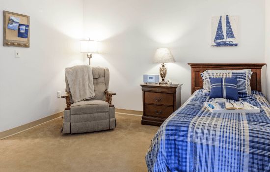 A cozy bedroom featuring a comfortable chair, nightstand with a lamp, and neatly made bed with checkered bedding.