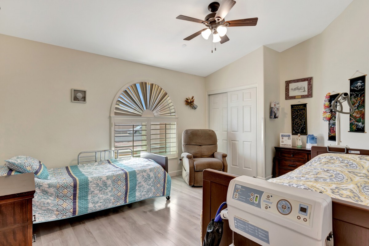 A cozy bedroom featuring twin beds, an armchair, and a ceiling fan, with sunlight streaming through the window shutters.