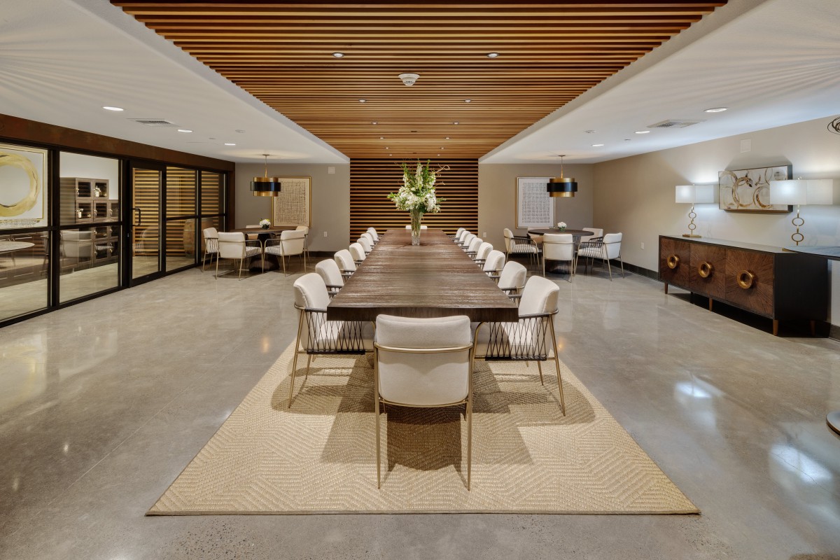 A modern conference room with a long wooden table surrounded by chairs, featuring elegant lighting and a floral centerpiece.