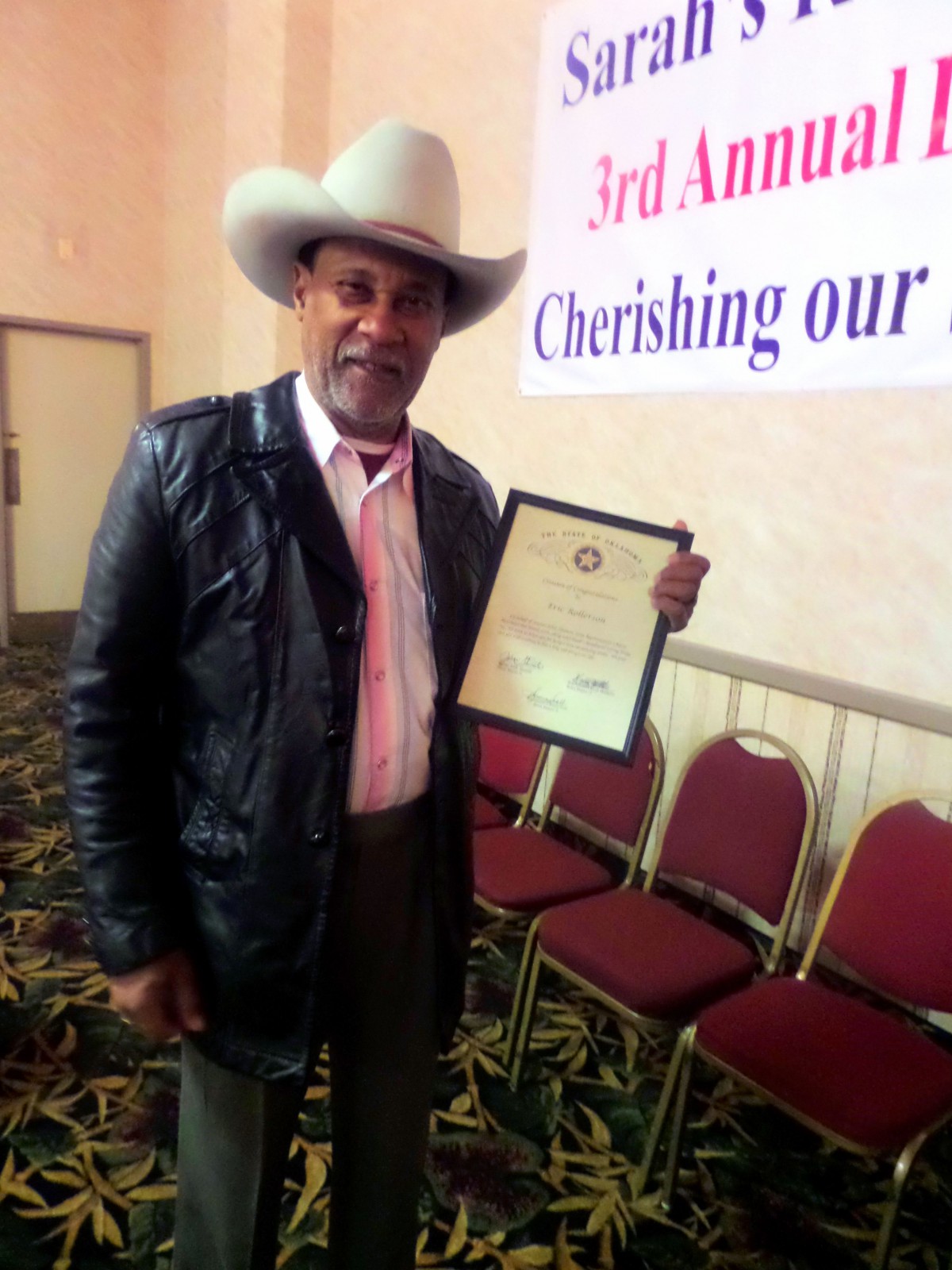 A person holding an award certificate at an event, wearing a cowboy hat and leather jacket.