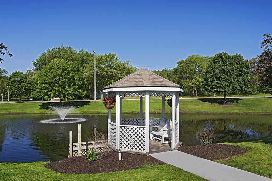 A scenic view of a gazebo by a pond with a fountain, surrounded by lush greenery.