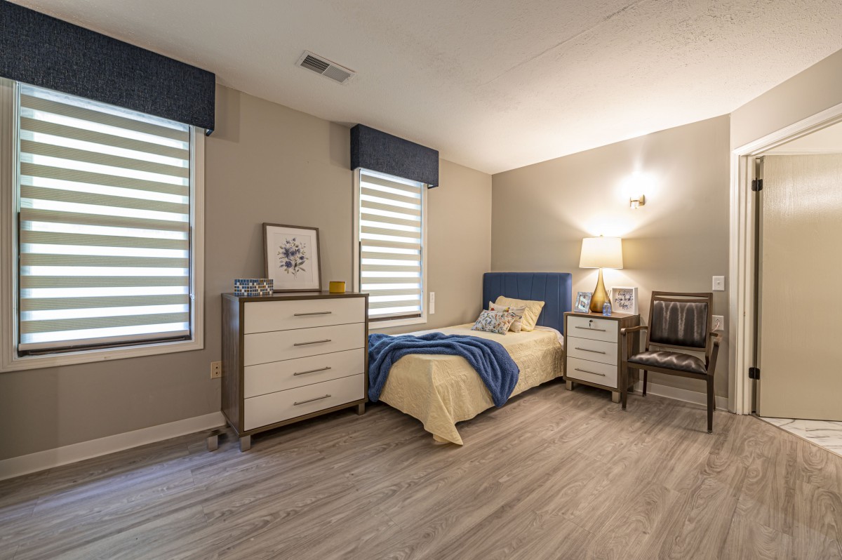 A modern bedroom featuring a single bed with a blue headboard, matching dressers, and striped window blinds.