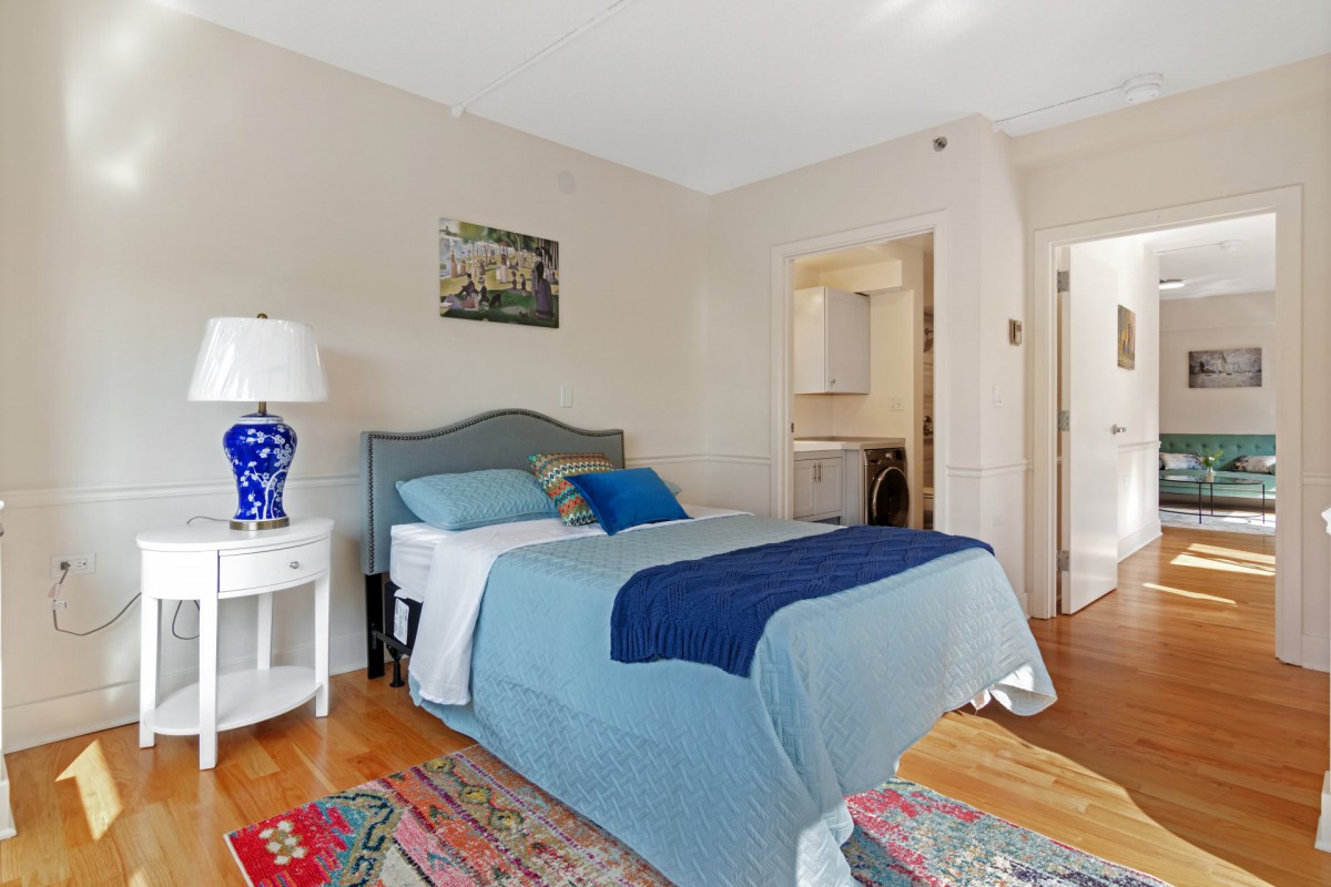 A cozy and modern bedroom featuring a bed with blue bedding, a white nightstand with a decorative lamp, and a colorful rug on the wooden floor.