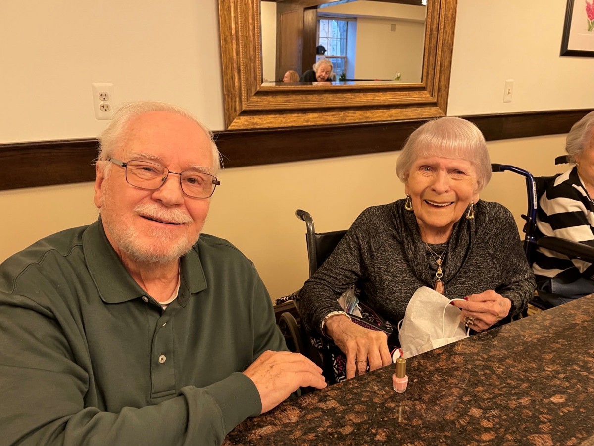 An elderly couple sitting together at a table, both smiling warmly.