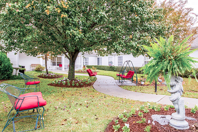 A serene garden setting with a pathway, surrounded by trees and equipped with red-cushioned patio furniture.