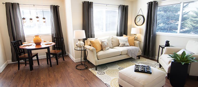 A cozy living room with a beige sofa adorned with mustard and cream throw pillows, flanked by lamps and dark curtains framing a bright window.