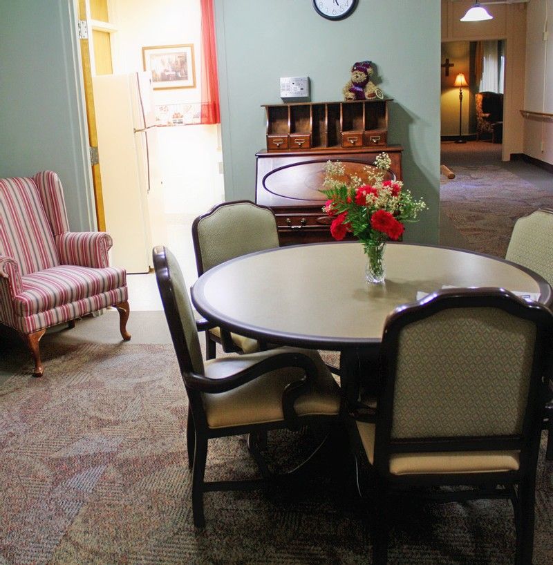 A cozy dining area with vintage furniture, including a round table and upholstered chairs, adorned with a bouquet of flowers.