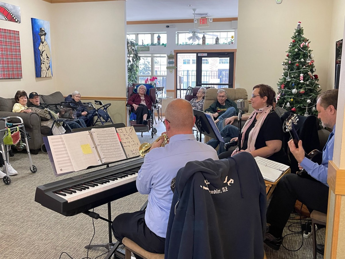A group of musicians performing indoors with a digital piano, in front of an audience and a Christmas tree.