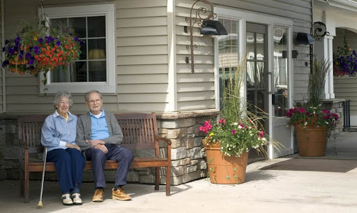 Two elderly individuals enjoying a sunny day on a bench outside a residential building.