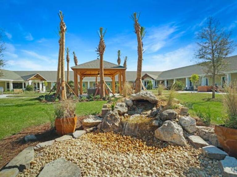 A serene garden featuring a gazebo surrounded by palm trees and a rock formation under a clear blue sky.