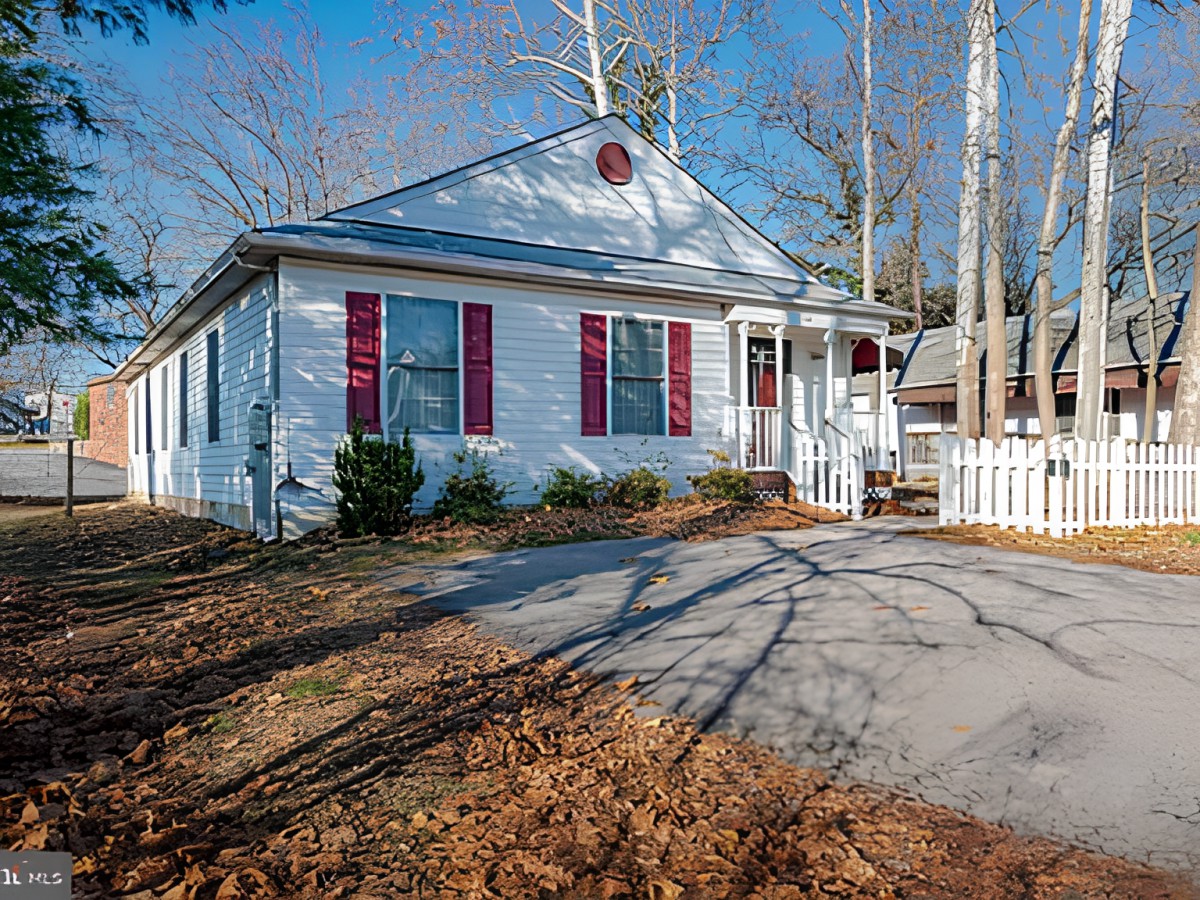 A cozy single-story house with a white exterior and red shutters, surrounded by trees and featuring a driveway.