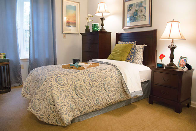 A cozy bedroom with a neatly made bed, complete with patterned bedding and decorative pillows. The room features artwork on the walls, a bedside lamp, and subtle decor elements.