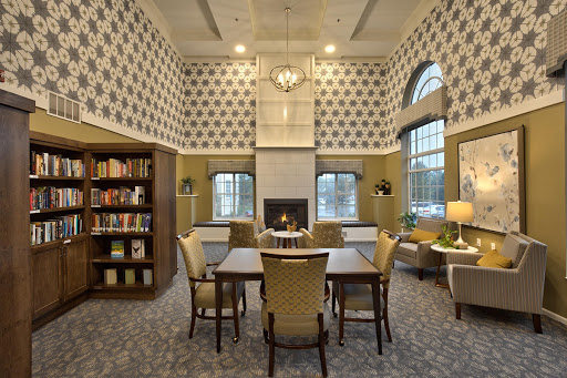 A cozy library space featuring comfortable seating and shelves filled with books, illuminated by natural light through large windows.