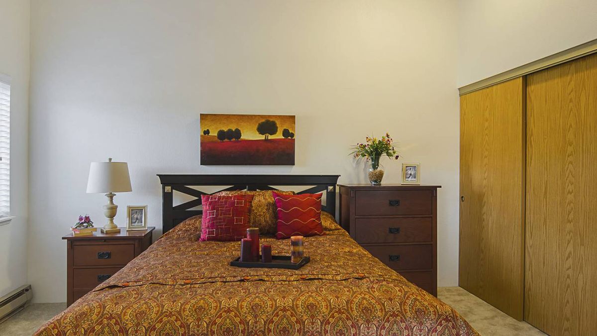 A cozy bedroom with a warm color palette, featuring a bed adorned with patterned bedding and vibrant pillows, complemented by a landscape painting above the headboard. A wooden dresser with a flower vase adds charm to the space.